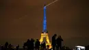 Para turis berada di sekitar Menara Eiffel diterangi dengan warna-warna khas Ukraina untuk menandai peringatan satu tahun invasi Rusia ke negara tersebut, di Paris, Kamis, 23 Januari 2023. Pada 24 Februari 2022 lalu Rusia menginvasi Ukraina dan menandai puncak eskalasi konflik kedua negara sejak 2014 silam. (AP Photo/Christophe Ena)