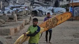 Dan, kawasan pantai di Kota Gaza menjadi salah satu sumber kebahagiaan berharga bagi warga Palestina yang ingin melepaskan diri dari tekanan situasi selama ini. Tampak dalam foto, warga Palestina, Khalil Abu Jayyab (depan) dan Tahseen Abu Assi membawa papan selancar mereka saat memasuki area sepanjang garis pantai di Kota Gaza, Minggu, 28 Desember 2025. (AP Photo/Jehad Alshrafi)