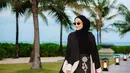 Zaskia Sungkar mengenakan outer panjang hitam dengan aksen putih di bagian bawah dipadukan inner dan kerudung warna senada. Serta bawahan celana panjang hitam bergaris dengan aksen apron dan bordiran bunga putih. [@zaskiasungkar15]