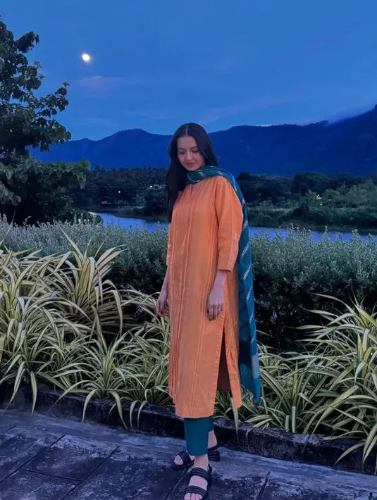 Raline memadukan pakaian model tunik orange dengan selendang warna biru bermotif garis putih. Serta mengenakan sandal hitam. [@ralineshah]