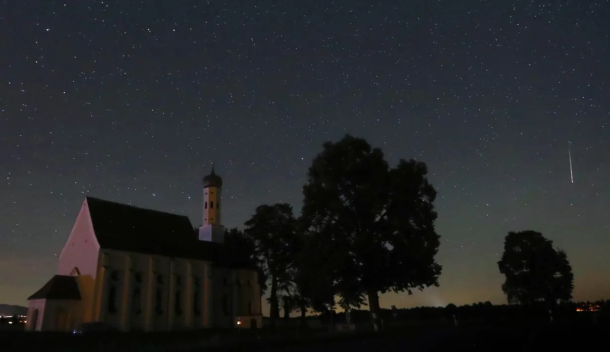 Sebuah meteor melintas di tengah langit malam selama hujan meteor tahunan Perseid di belakang gereja ziarah Sankt Coloman, barat daya Jerman, Minggu (12/8). Puncak hujan meteor terjadi pada 11-12 Agustus dan 12-13 Agustus. (KARL-JOSEF HILDENBRAND/DPA/AFP)