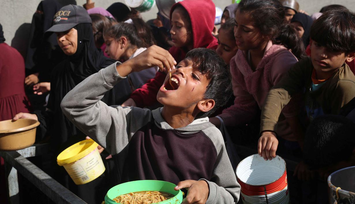 Hingga Minggu 23 November 2025, antrean warga untuk mendapatkan bantuan makanan masih terlihat di sejumlah titik pengungsian, salah satunya di Nuseirat, pusat Jalur Gaza. Tampak dalam foto, seorang anak laki-laki Palestina yang terlantar sedang makan pasta setelah menerima porsi makanan di sebuah tempat penampungan yang selama ini dihuni banyak keluarga, di Nuseirat, pusat Jalur Gaza, pada 23 November 2025. (EYAD BABA/AFP)