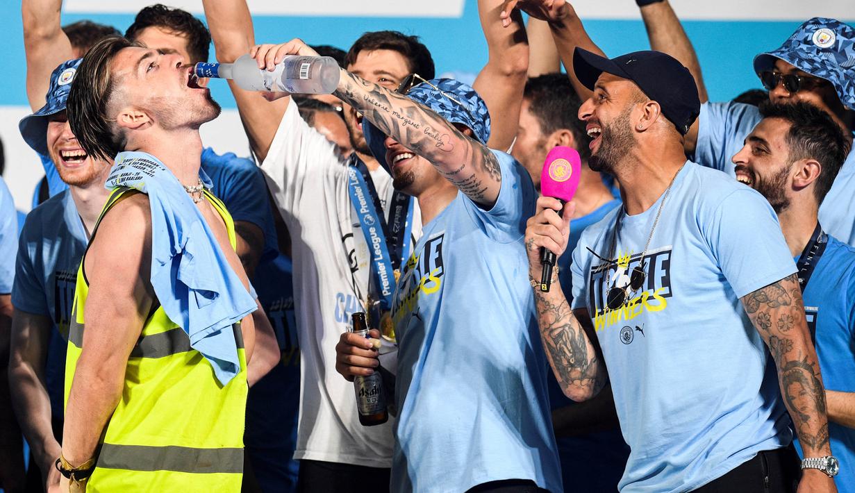 Pemain Manchester City, Jack Grealish (kiri), menggunakan rompi proyek disaat pemain lainnya memakai seragam dalam perayaan Trebele Winners di kota Manchester, Selasa (13/6/2023) dini hari WIB. Salah satu rekan satu timnya, Kalvin Phillips (tengah) bahkan menyuguhkan minuman beralkhol langsung ke mulut Grealish. (AFP/Oli Scarff)