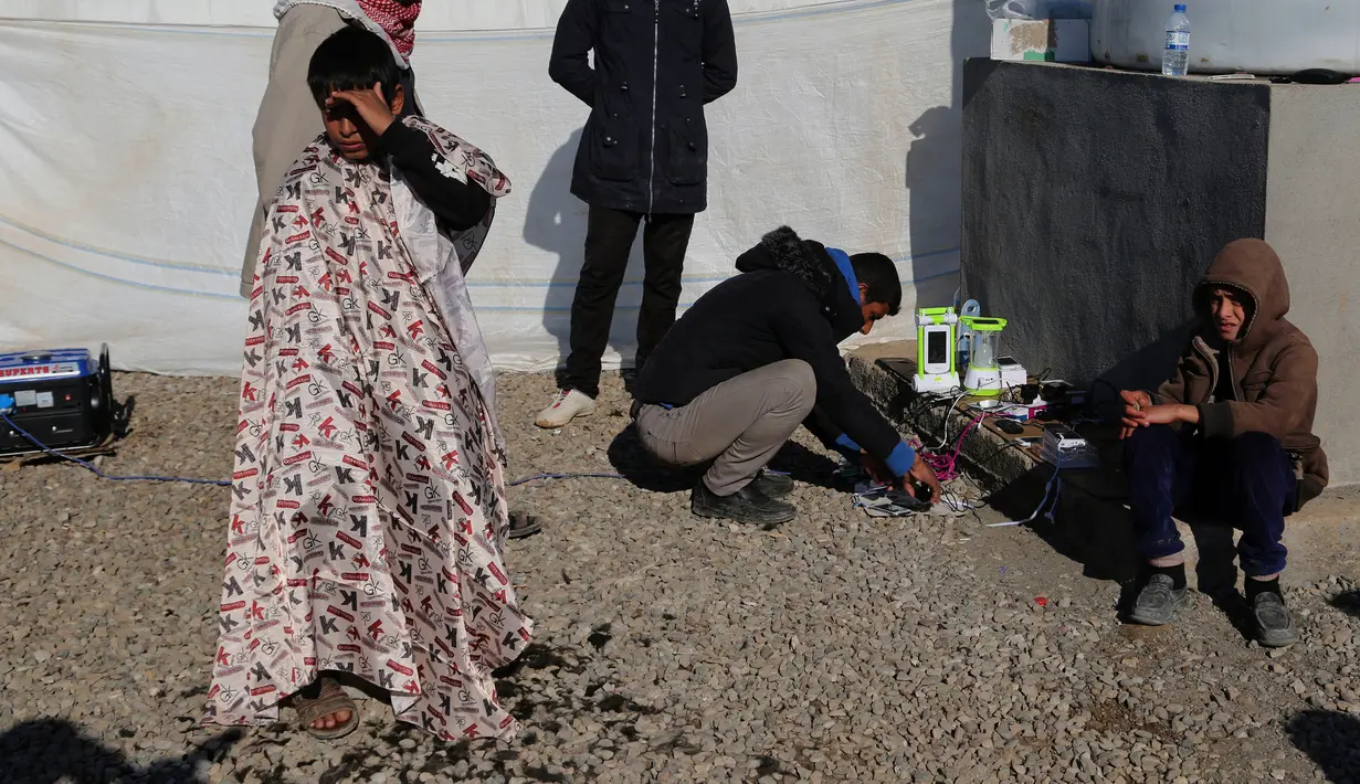 Seorang anak menunggu untuk memotong rambutnya sementara pengungsi lainnya terlihat mengisi baterai ponselnya untuk berkomunikasi dengan rekannya di kamp pengungsian Sewdinan, Khazer, Irak, 3 Januari 2017. (AP Photo / Khalid Mohammed)
