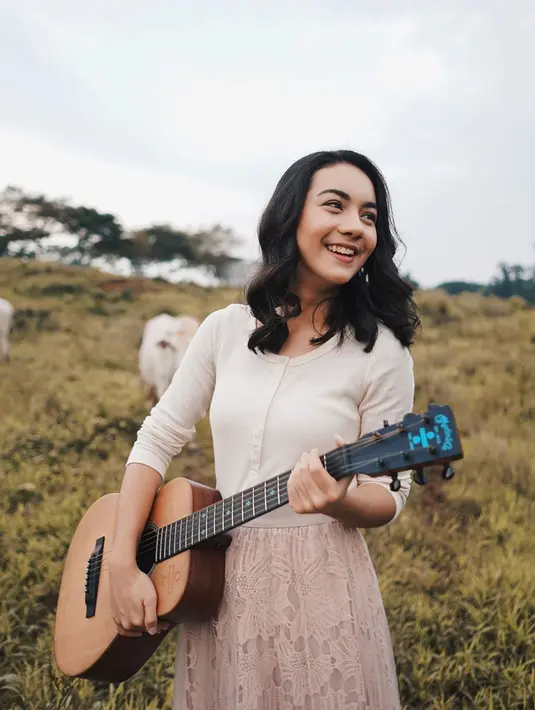 Seperti ketika ia sedang menikmati ladang rumput dengan sapi-sapi. Ersya tampak cantik dengan atasan putih dan rok panjang berwarna merah muda serta memegang gitar. (Liputan6.com/IG/@ersyaurel)