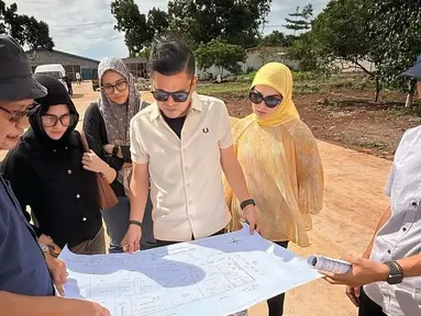 Dalam momen yang dibagikan, Haldy terlihat memegang cetak biru proyek sambil berdiskusi dengan tim di lokasi yang masih dalam tahap awal pembangunan. Irish Bella, mengenakan busana kuning dan hijab senada, setia mendampingi suaminya. Pemandangan area terbuka dan tanah merah menunjukkan proyek yang tengah digarap secara serius. (Liputan6.com/IG/@haldy_sabri)