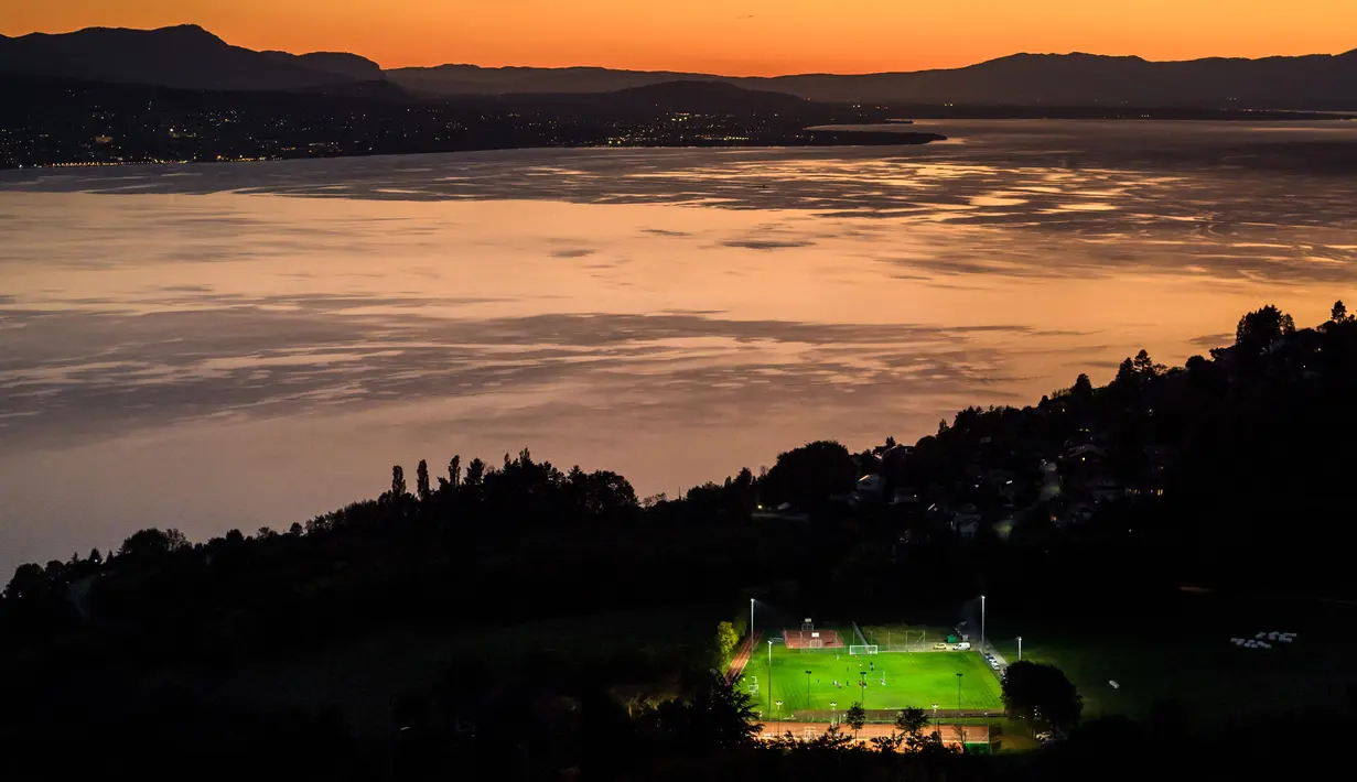 <p>Lapangan sepak bola yang berada di kawasan Mont de Gourze, Swiss barat. Keindahan suasana di sekitar lapangan semakin menjunjang berkat lampu sorot yang di latari dengan danau Jenewa yang begitu memukau. (AFP/Fabrice Coffrini)</p>