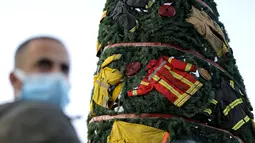 Seorang pria berada di dekat pohon Natal yang dihiasi pakaian dan peralatan milik petugas pemadam kebakaran yang tewas dalam ledakan di Pelabuhan Beirut, Lebanon (20/12/2020). Pohon Natal itu dihias oleh seorang seniman Lebanon bernama Hayat Nazer. (Xinhua/Bilal Jawich)
