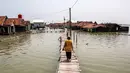 Sementara, warga yang bertahan beradaptasi dengan meninggikan rumah, membangun jembatan kayu secara swadaya, atau mendirikan rumah apung. Tampak dalam foto, seorang wanita berjalan melintasi jembatan melewati rumah-rumah yang sebagian terendam air laut akibat banjir pasang di desa Timbulsloko, Demak, Jawa Tengah pada Sabtu 18 April 2026. (DEVI RAHMAN/AFP)