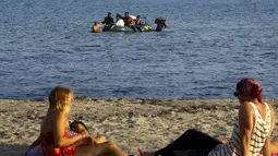Wisatawan berbaring di pantai melihat para imigran dan pengungsi dari Suriah dan Afrika tiba saat berada diperahu usai melintasi Laut Aegea antara Turki dan Yunani, (8/8/2015). (REUTERS/Yannis Behrakis)