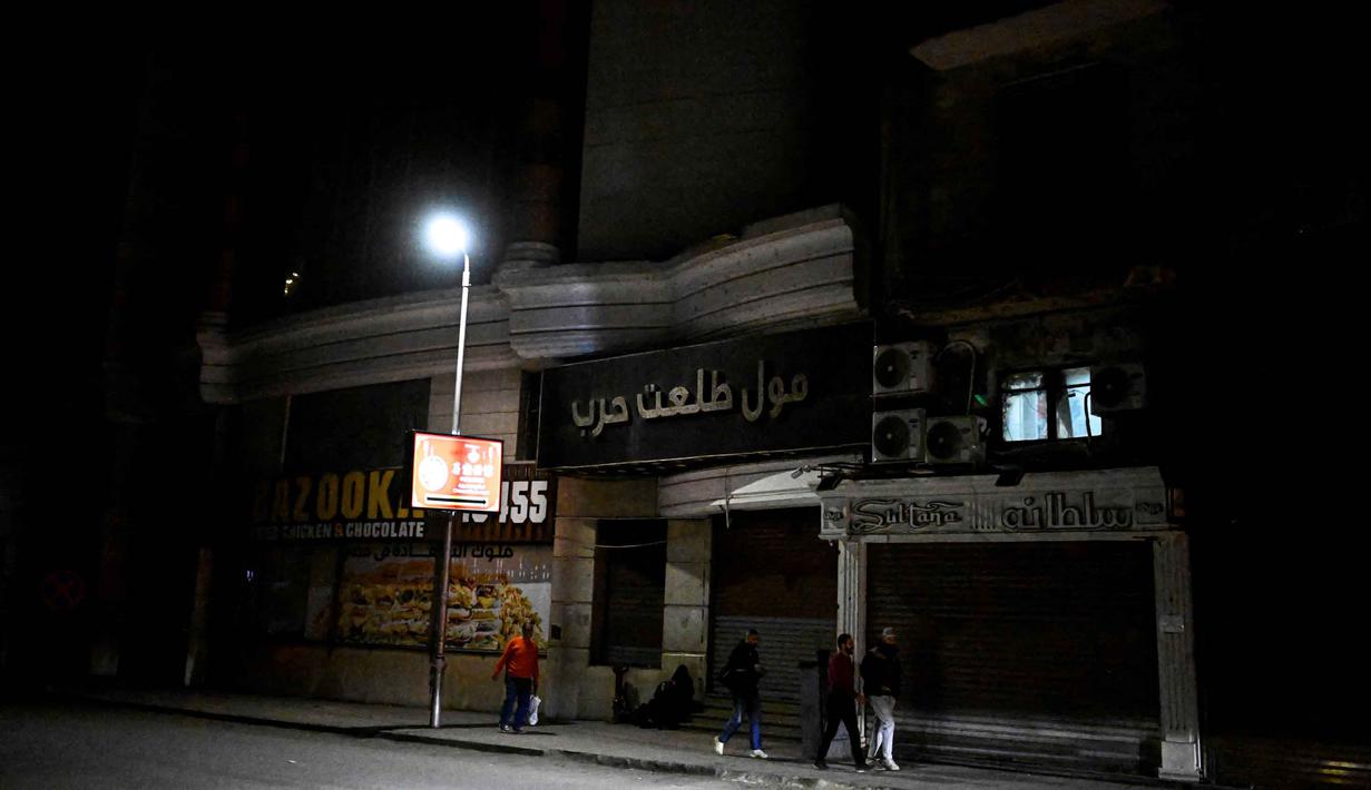 Kebijakan ini merupakan bentuk respons keras Pemerintah Mesir atas dampak yang ditimbulkan akibat perang antara Amerika Serikat (AS)-Israel dengan Iran. Tampak dalam foto, orang-orang berjalan melewati toko-toko yang tutup lebih awal karena jam malam yang diberlakukan pemerintah untuk mengurangi biaya energi di pusat kota Kairo, Mesir, pada Kamis 2 April 2026. (Khaled DESOUKI/AFP)