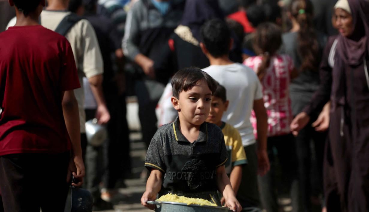 Seorang anak laki-laki Palestina yang sedang bermain membawa sepanci makanan yang ia terima dari sebuah tempat penampungan tempat tinggal keluarga-keluarga di Nuseirat, pusat Jalur Gaza, pada 2 November 2025. PBB juga menyerukan agar semua titik perbatasan dibuka dan lebih banyak lembaga kemanusiaan diberi izin membawa bantuan ke Gaza. (EYAD BABA/AFP)