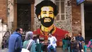 Foto file pada 4 April 2018 menunjukkan orang-orang duduk di sebuah kafe di pusat kota Kairo dengan latar belakang lukisan mural yang menggambarkan striker Liverpool, Mohamed Salah. Federasi Sepak Bola Mesir melaporkan, Jumat (13/11/2020), Mohamed Salah dinyatakan positif Covid-19. (Amir MAKAR/AFP)