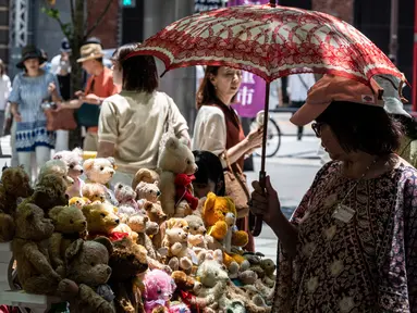 Seorang perempuan (kanan) menggunakan payung untuk berlindung dari teriknya sinar matahari saat melihat-lihat boneka beruang yang dijual di pasar barang antik di pusat kota Tokyo, Jepang, Senin (17/7/2023). (Richard A. Brooks / AFP)