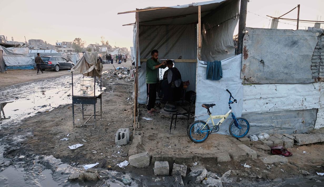 Untuk diketahui, Perserikatan Bangsa-Bangsa (PBB) memperkirakan sekitar 92 persen dari seluruh bangunan tempat tinggal, atau kurang lebih 436.000 hunian di Gaza, termasuk Jabalia telah rusak atau hancur sejak dimulainya konflik pada Oktober 2023 lalu. Tampak dalam foto, seorang tukang cukur memotong rambut seorang pria di dalam tenda darurat di kamp pengungsian di Jabalia, di bagian utara Jalur Gaza, Minggu 7 Desember 2025. (AP Photo/Jehad Alshrafi)