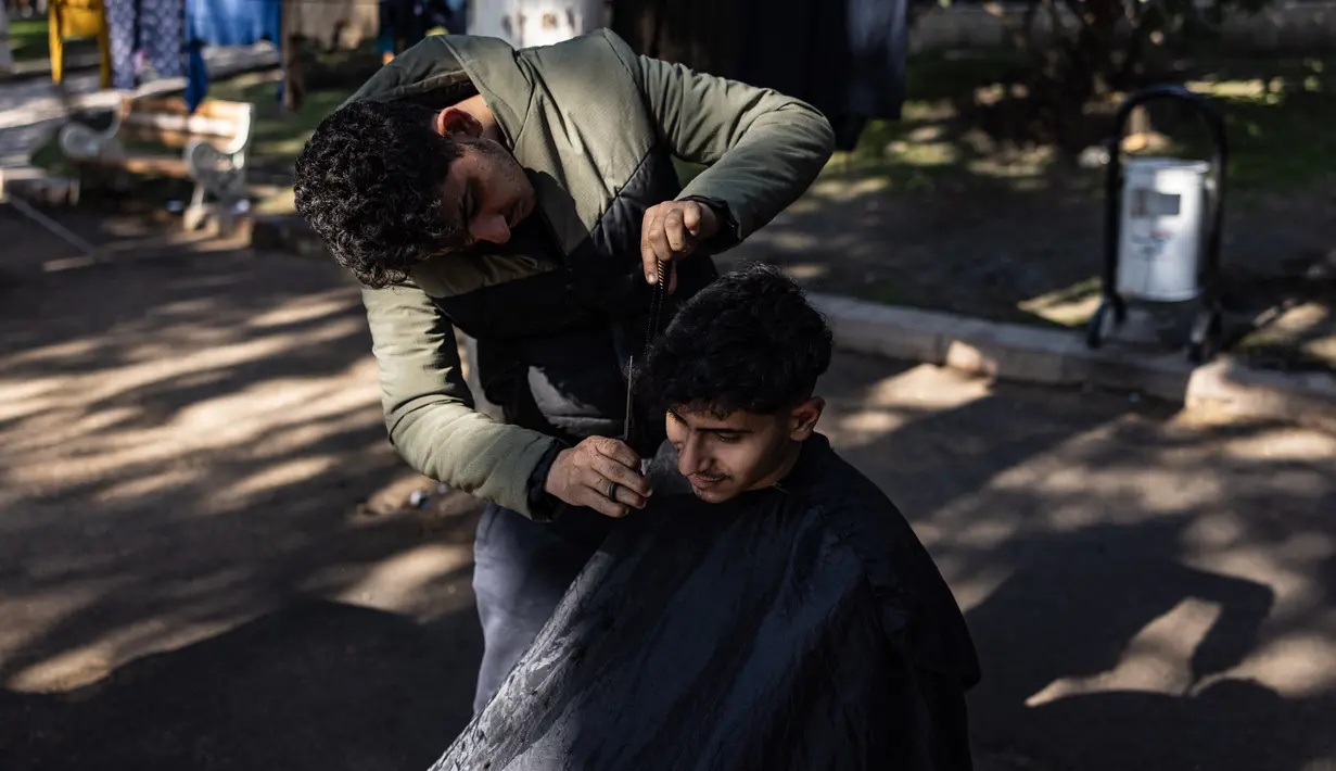 Mohammed al-Hamo (kiri) memotong rambut saudara laki-lakinya, Sobhi di depan tenda mereka di kamp darurat pengungsi korban gempa Turki dan Suriah di kota Antakya pada 19 Februari 2023. Mohammed al-Hamo tak pernah membayangkan akan menggunakan keterampilannya mencukur rambut akan digunakan di tenda pengungsi korban gempa Turki dan Suriah. (Sameer Al-DOUMY / AFP)