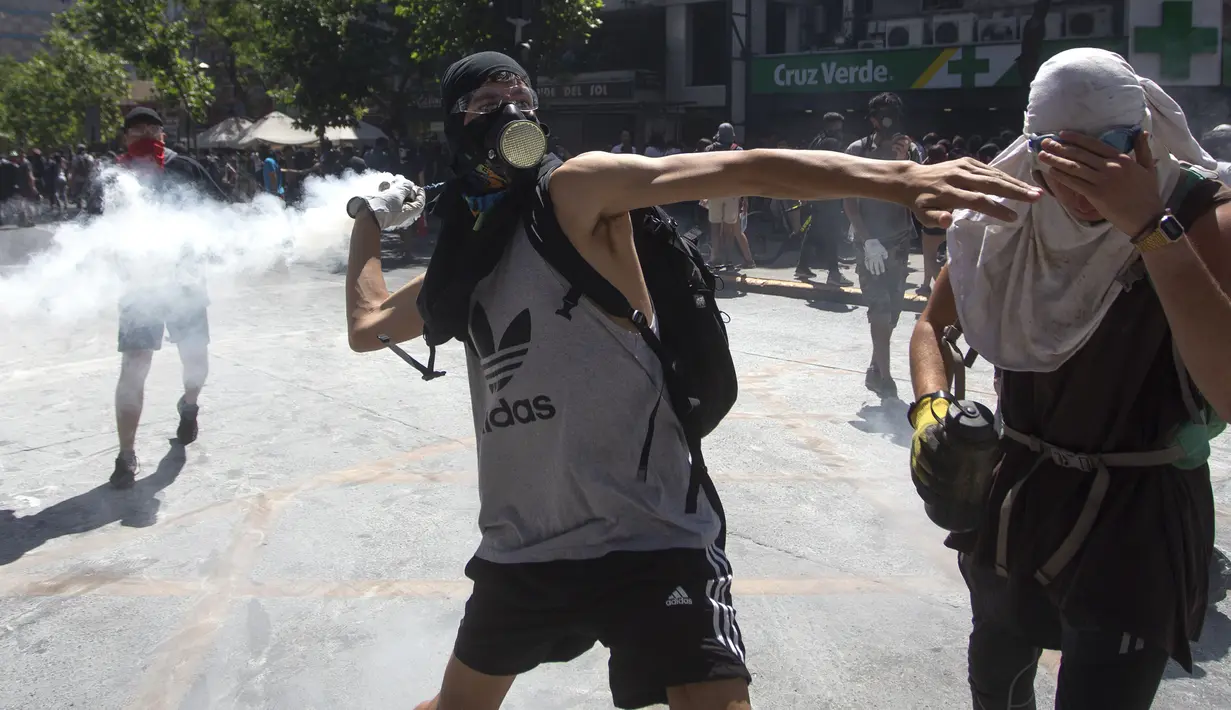 Seorang demonstran melempar tabung gas air mata ke polisi anti huru hara selama protes terhadap kebijakan ekonomi pemerintah di dekat Costanera Center (kompleks bisnis dan komersial) di Santiago (6/11/2019). (AFP Photo/Claudio Reyes)