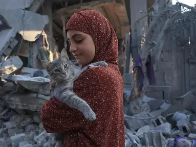 Seorang anak perempuan Palestina menggendong seekor kucing saat dia memeriksa kerusakan setelah pemboman Israel di Rafah di Jalur Gaza selatan, pada 18 Oktober 2023. (SAID KHATIB/AFP)