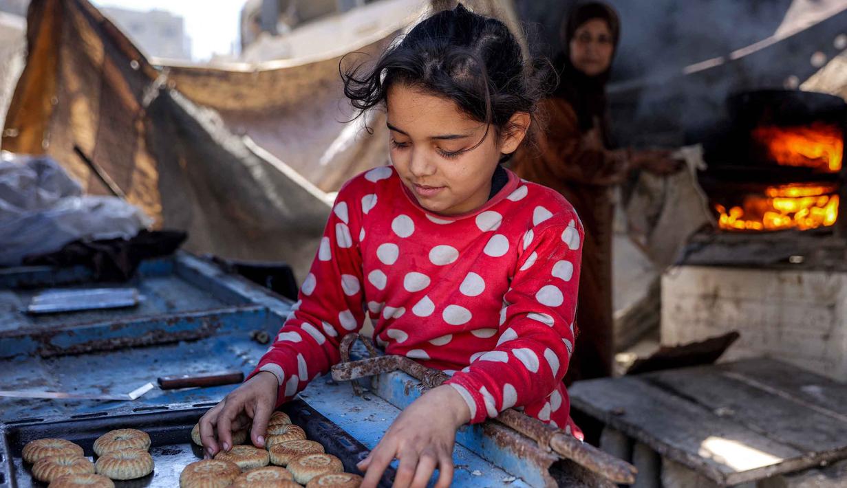 Meski secara bentuk kue Kaak menyerupai roti, namun teksturnya yang renyah di luar dan empuk di dalam. Tampak dalam foto, seorang gadis memilah-milah biskuit tradisional "kaak" yang baru dipanggang di atas nampan, yang dibuat menjelang Idul Fitri, hari raya yang menandai berakhirnya bulan suci puasa Ramadhan, di Kota Gaza pada 16 Maret 2026. (Omar AL-QATTAA/AFP)