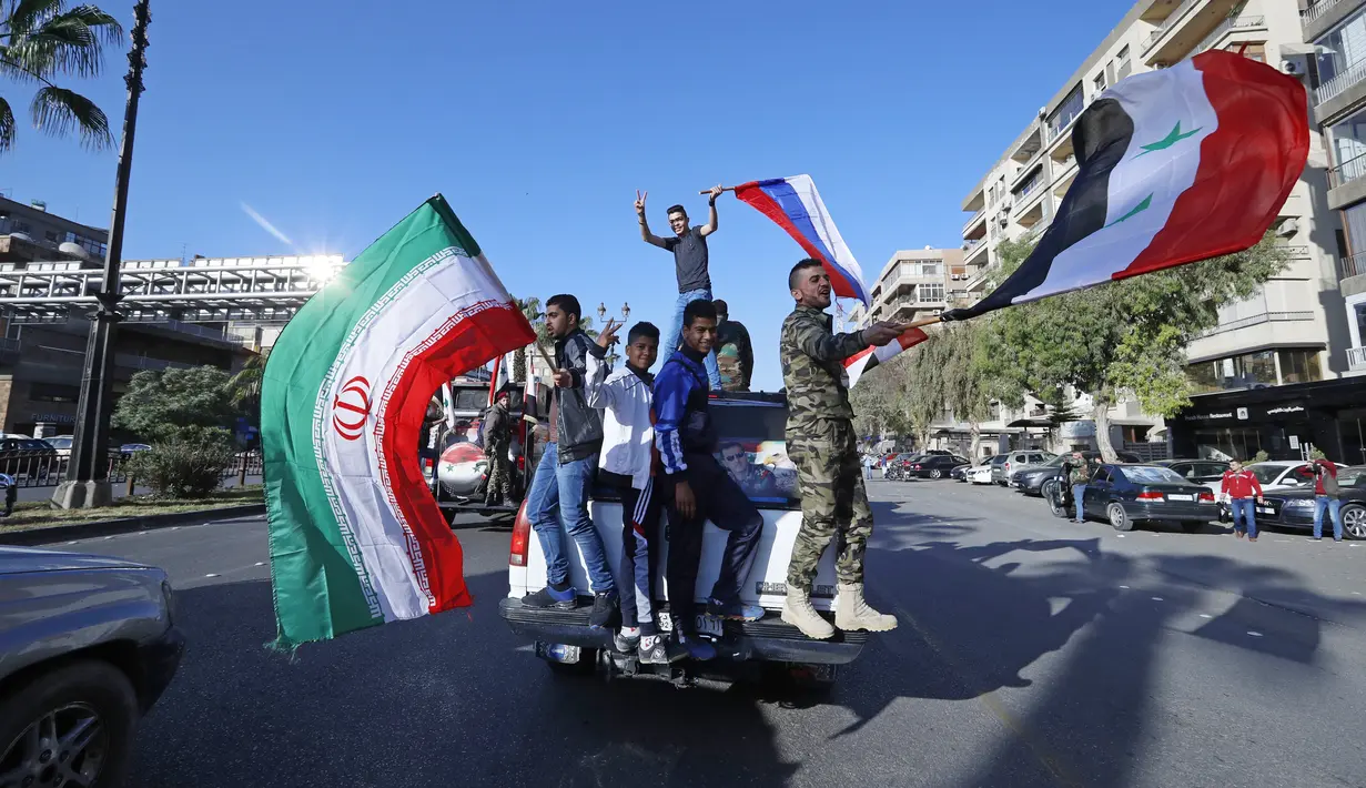 Pendukung pemerintah mengibarkan bendera Suriah, Iran, dan Rusia saat berdemonstrasi di Damaskus, Suriah, 14 April 2018. Suriah tengah mengalami gejolak krisis ekonomi dengan nilai tukar mata uang yang merosot dalam beberapa hari terakhir. (AP Photo/Hassan Ammar, File)