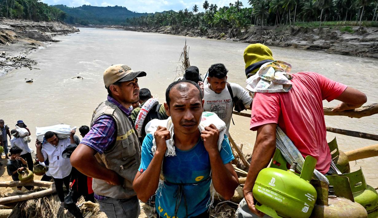 Tumpukan puing kayu menjadi solusi sementara sekaligus jalur alternatif bagi warga untuk melanjutkan aktivitas harian dan mengambil bantuan logistik. Tampak dalam foto, warga menyeberangi sungai di jembatan yang baru dibangun yang menghubungkan Aceh dan Provinsi Sumatera Utara setelah hancur akibat banjir bandang di Sungai Peusangan, Distrik Bireuen, Provinsi Aceh, Selasa 9 Desember 2025. (CHAIDEER MAHYUDDIN/AFP)