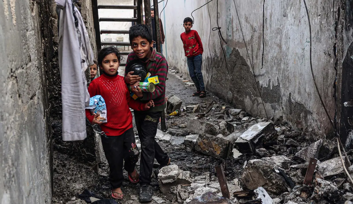 Anak-anak membawa barang-barang makanan yang diselamatkan dari rumah mereka yang rusak saat mereka menemukan jalan melalui gang yang tertutup puing-puing, setelah pengeboman Israel di Rafah, Jalur Gaza selatan pada 25 Februari 2024. (SAID KHATIB/AFP)