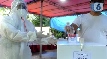 Petugas Kelompok Penyelenggara Pemungutan Suara (KPPS) dengan memakai baju hazmat membantu pemilih memasukkan kertas suara usai menggunakan hak pilihnya dalam Pilkada Tangerang Selatan 2020 di TPS 46, Paku Jaya, Serpong Utara, Rabu (9/12/2020). (merdeka.com/Arie Basuki)