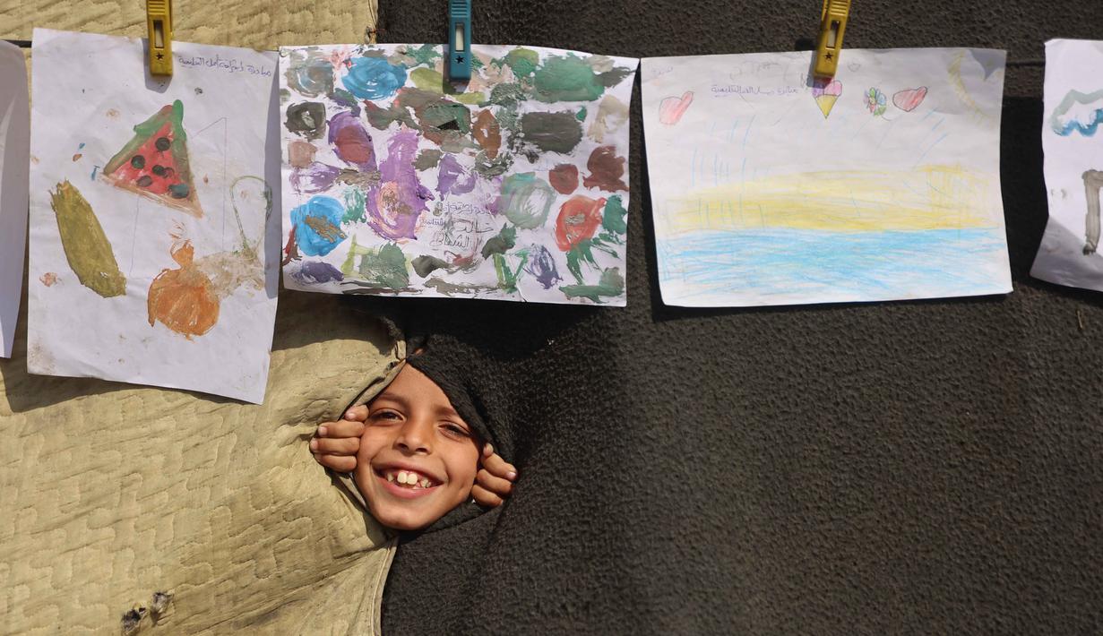 Seorang anak melihat melalui lubang di selimut selama pameran yang menampilkan koleksi gambar oleh anak-anak dan perempuan di sebuah sekolah yang diubah menjadi tempat penampungan bagi pengungsi Palestina di kawasan Al-Rimal, Kota Gaza, pada 5 November 2025. (Omar AL-QATTAA/AFP)