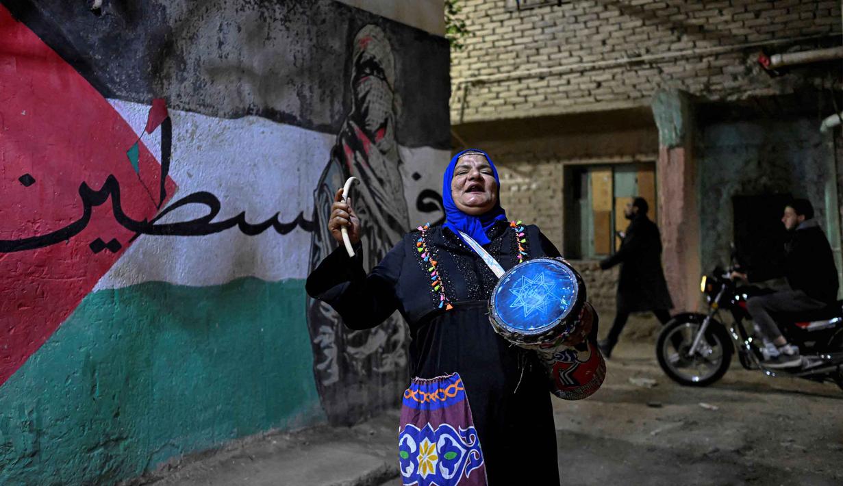 : Saat ini, meski teknologi seperti alarm sudah umum digunakan, Musaharati tetap dipertahankan sebagai bagian dari warisan budaya yang mempererat hubungan warga. Tampak dalam foto, Dalal, seorang wanita Mesir 'musaharati', berdiri di depan grafiti yang menggambarkan bendera Palestina di sepanjang jalan sambil memukul gendang dan menyerukan umat Muslim untuk bangun dan makan sahur selama bulan suci Ramadan di distrik Hadayek el-Maadi, selatan Kairo pada Minggu 1 Maret 2026. (Khaled DESOUKI/AFP)
