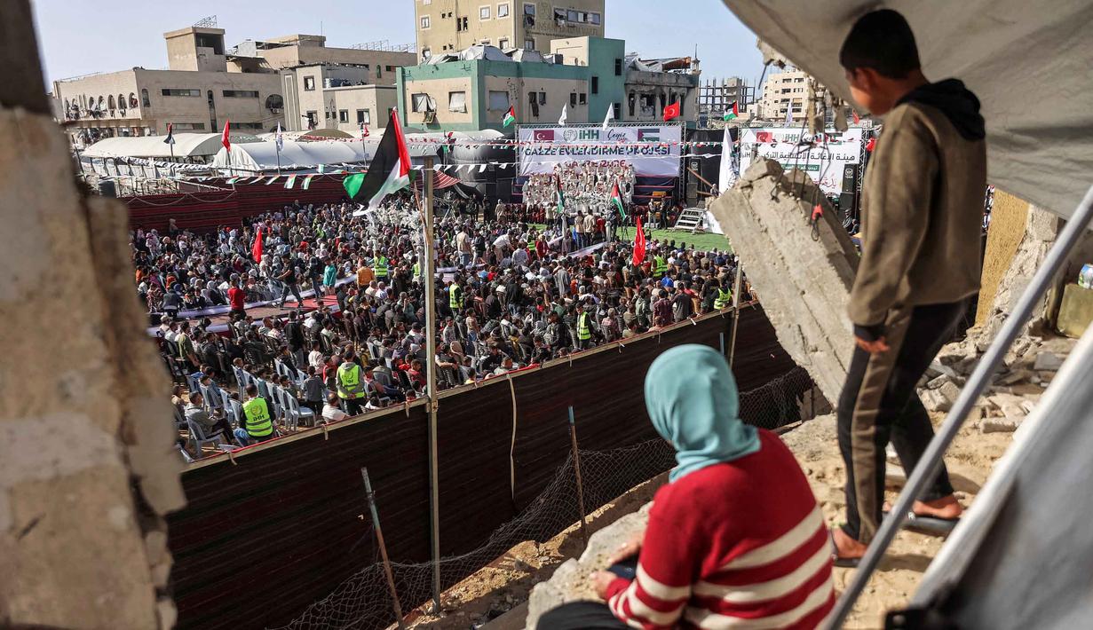 Kegiatan ini digelar di tengah bayang-bayang blokade dan kondisi kemanusiaan yang masih berlangsung di Gaza. Tampak dalam foto, seorang anak laki-laki dan perempuan mengamati dari sebuah bangunan yang rusak parah saat orang-orang berkumpul selama perayaan pernikahan massal untuk para pengantin Palestina yang diselenggarakan oleh Yayasan Bantuan Kemanusiaan Turki (IHH) di Kota Gaza pada 11 Mei 2026. (Omar AL-QATTAA/AFP)