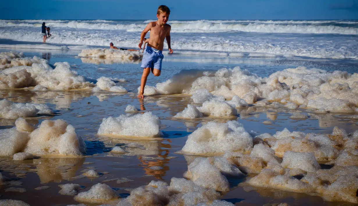 Seorang anak bermain di antara busa pantai setelah siklon tropis di Currumbin Beach, Gold Coast, Australia pada 15 Desember 2020. Hujan lebat dan angin kencang telah melanda kawasan perbatasan padat penduduk antara New South Wales dan Queensland selama tiga hari. (Photo by Patrick HAMILTON / AFP)