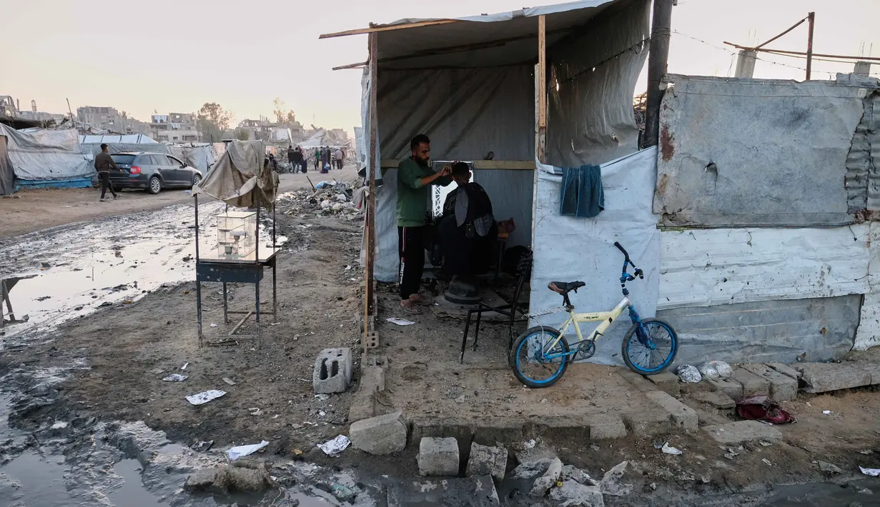 Untuk diketahui, Perserikatan Bangsa-Bangsa (PBB) memperkirakan sekitar 92 persen dari seluruh bangunan tempat tinggal, atau kurang lebih 436.000 hunian di Gaza, termasuk Jabalia telah rusak atau hancur sejak dimulainya konflik pada Oktober 2023 lalu. Tampak dalam foto, seorang tukang cukur memotong rambut seorang pria di dalam tenda darurat di kamp pengungsian di Jabalia, di bagian utara Jalur Gaza, Minggu 7 Desember 2025. (AP Photo/Jehad Alshrafi)