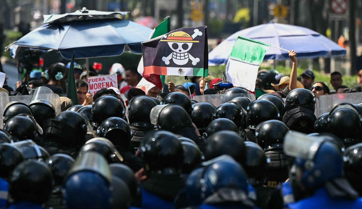Unjuk rasa ini untuk memprotes meningkatnya angka kejahatan, korupsi, dan impunitas di Meksiko. Tampak dalam foto, seorang pengunjuk rasa memegang bendera berlogo manga populer Jepang, One Piece, simbol yang diadopsi oleh gerakan protes Gen Z di seluruh dunia, di depan barisan polisi di sepanjang Reforma Avenue dalam sebuah demonstrasi yang dilakukan oleh anak muda Meksiko terkait dengan gelombang protes global Generasi Z terhadap pemerintahan Presiden Meksiko, Claudia Sheinbaum, di Mexico City, pada 20 November 2025. (Yuri CORTEZ/AFP)