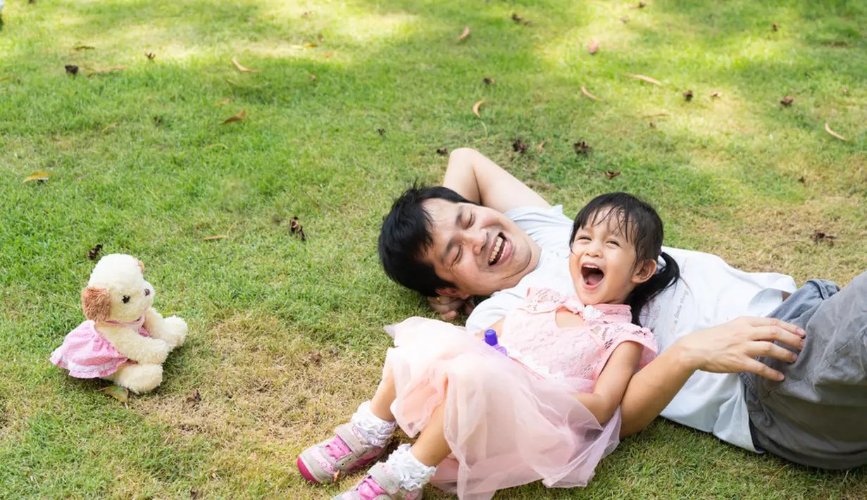 Potret ayah dan anak berbaring dan tertawa bersama dengan penuh kebahagiaan saat bermain di atas hamparan rumput yang hijau. Gemes sekali, ya, melihatnya. (SUKJAI PHOTO/ Shutterstock.com)