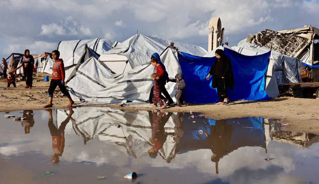 Warga Palestina yang mengungsi berjalan melewati genangan air di dekat tenda-tenda penampungan saat wilayah tersebut dilanda hujan dan kondisi musim dingin yang dingin, di Kota Gaza pada 28 Desember 2025. Cuaca ekstrem serta musim dingin yang ekstrem di Jalur Gaza semakin memperparah krisis kemanusiaan di tengah keterbatasan tempat berlindung yang aman. (Omar AL-QATTAA/AFP)