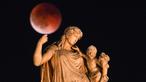 Foto ini menampilkan bulan purnama yang juga dikenal sebagai “Blood Moon” selama gerhana bulan, dengan patung dewi Yunani kuno Irene yang memegang seorang anak di latar depan, di pusat Athena, Yunani pada 7 September 2025. (Angelos TZORTZINIS/AFP)