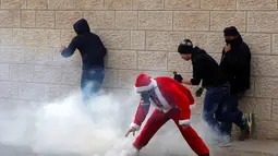 Seorang pengunjuk rasa berkostum Santa Claus berusaha mengambil gas air mata saat aksi di desa Tepi Barat Bilin dekat Ramallah, Jumat (23/12). Mereka menentang pembangunan tembok pembatas di Israel. (REUTERS/Mussa Issa Qawasma)