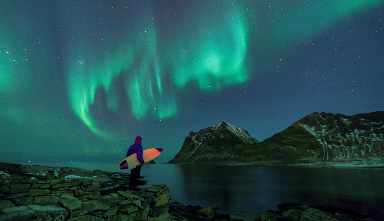 Seorang peselancar menatap Aurora sebelum beraksi di Lofoten Islands, Norwegia, (9/3/2018). Suhu udara minus 13°C dan suhu diatas air sekitar 4°C. (AFP/Oliver Morin)