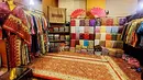 Busana songket berwarna merah marun dengan motif segitiga dan garis geometris emas, serta paduan kain bercorak diagonal yang kaya warna dan deretan kain songket lipat dan pajangan kain utuh yang memamerkan variasi motif tradisional seperti flora, geometris, dan ornamen khas tenun. Palet warnanya sangat beragam, mulai dari emas, perak, ungu, merah, biru, hingga kombinasi warna kontras, semuanya memancarkan kilau benang logam yang menjadi ciri khas kemewahan kain songket. (Foto: instagram@/zainalsongket)