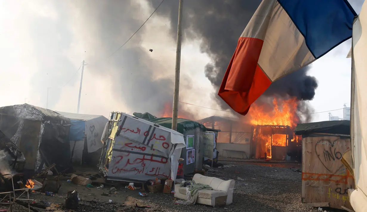 Sebuah bendera Prancis berada di dekat Kobaran api yang membakar sebuah kamp pengungsian imigran selama pembongkaran kamp pengungsian imigran "Jungle", di kota pelabuhan Calais, Prancis (26/10). (REUTERS/Pascal Rossignol)