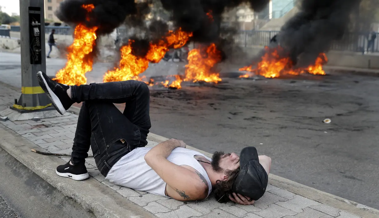 Pengunjuk rasa berbaring di tanah saat ia beristirahat dekat ban yang terbakar di jalan raya utama yang mengarah ke Bandara Internasional Beirut selama protes terhadap kenaikan harga barang-barang konsumen dan jatuhnya mata uang lokal di Beirut, Lebanon, Senin (29/11/2021). (AP Photo/Hussein Malla)