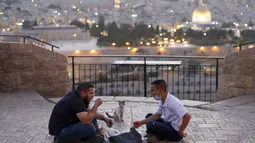 Dua pria Pelestina berbuka puasa selama bulan suci Ramadan dengan latar belakang Kota Tua Yerusalem dan kompleks Masjid al-Aqsa yang ditutup selama krisis pandemi coronavirus yang baru di Bukit Zaitun (19/5/2020). (AFP/Ahmad Gharabli)