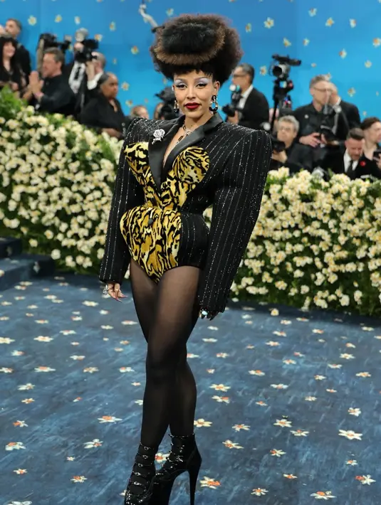 Doja Cat mengenakan bodysuit custom rancangan Marc Jacobs yang terinspirasi dari gaya power dressing tahun 1980-an. Bodysuit menampilkan bahu lebar dengan padding dramatis, motif pinstripe klasik, dan detail bustier bermotif leopard print berwarna kuning cerah yang berpuncak pada cone bra khas. Doja melengkapi penampilannya dengan stocking hitam transparan, sepatu platform bergaya wingtip, serta aksesori seperti clutch leopard, perhiasan vintage, dan bros berbentuk mawar. Rambutnya ditata dengan volume besar, menambah kesan retro yang kuat. [Marc Jacobs ]