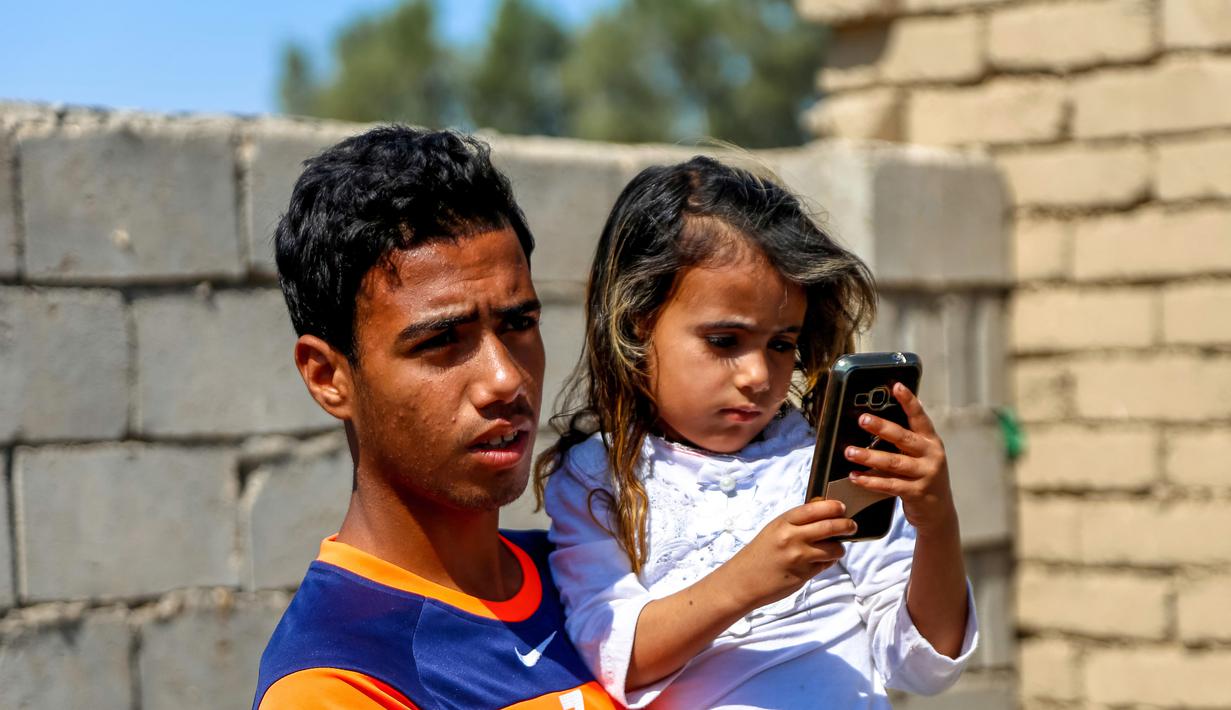 Houra bermain ponsel saat digendong saudaranya di desa Wahed Haziran, Provinsi Diwaniya, Irak, (17/3). Penyakit langka ini membuat Houra rentan terserang kanker kulit paling berbahaya, yakni melanoma. (AFP Photo/Haidar Hamdani)