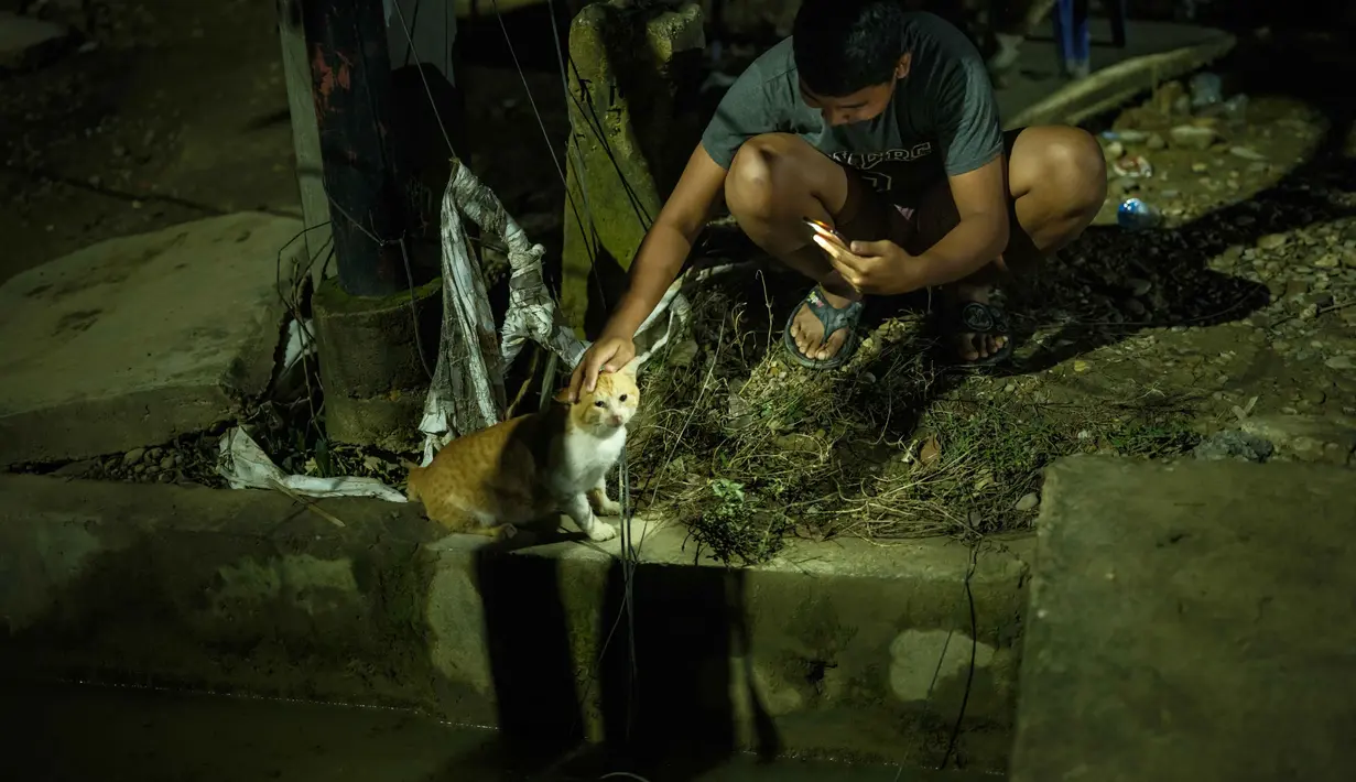 Warga yang mengungsi terpaksa menggunakan lampu minyak darurat atau pelita untuk penerangan. Tampak dalam foto, seorang siswa SMA berusia 16 tahun, yang kehilangan rumahnya akibat banjir bandang sekitar tiga minggu sebelumnya, merekam seekor kucing dengan ponselnya di depan tempat tinggal sementara yang ia bangun di Aceh Tamiang, pada Selasa 16 Desember 2025. (Yasuyoshi Chiba/AFP)