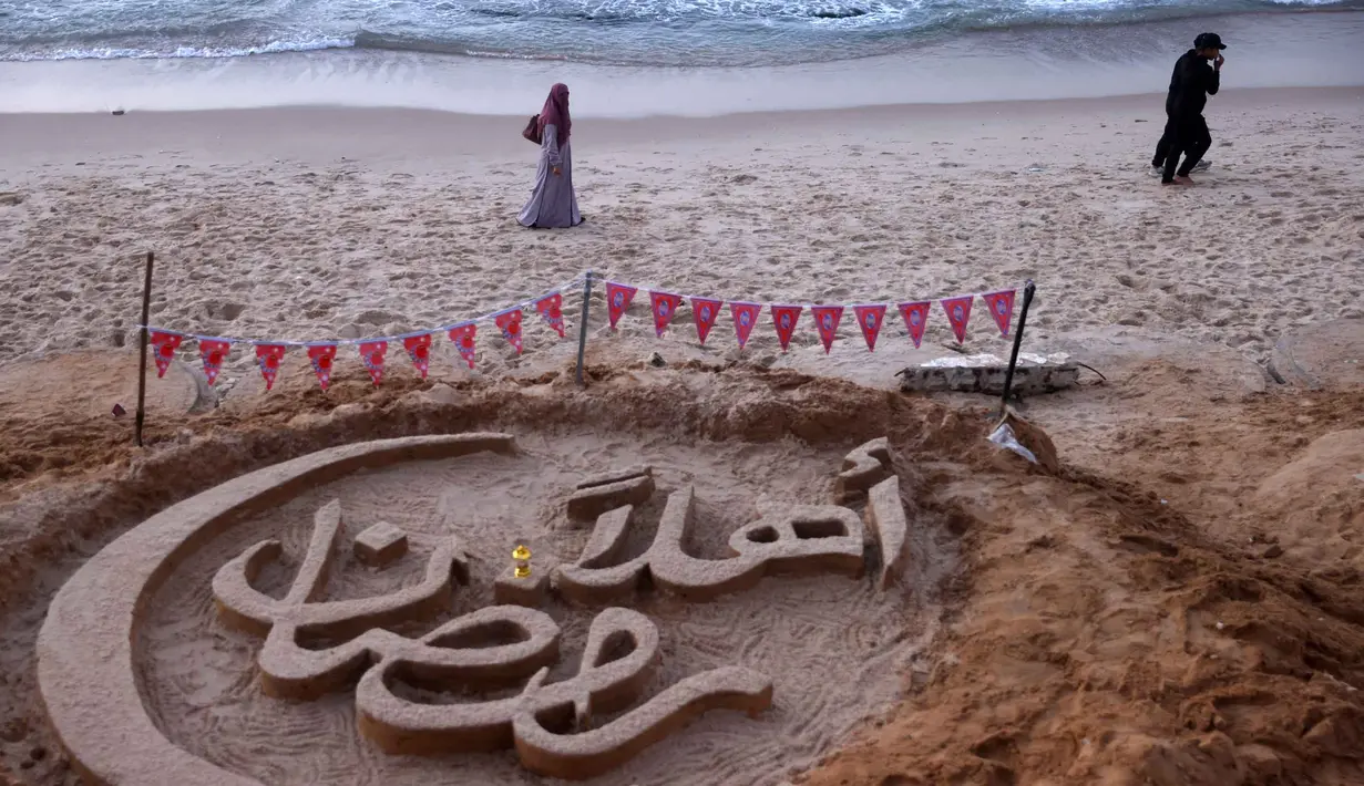 Kegiatan menjadi upaya menghadirkan keceriaan dan nuansa perayaan Ramadan di tengah kehancuran akibat konflik. Tampak dalam foto, orang-orang berjalan melewati patung pasir dengan pesan ucapan "Selamat Datang, Ramadan" yang dibuat oleh seniman Palestina, Yazeed Abu Jarad, di sepanjang pantai di Khan Younis, selatan Gaza pada 17 Februari 2026. (Bashar Taleb/AFP)