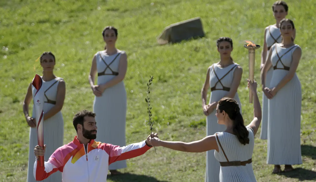Aktris Yunani Xanthi Georgiou yang berperan sebagai High Priestess menyerahkan ranting zaitun kepada pembawa obor pertama pembalap ski alpine Yunani Ioannis Antoniou saat penyalaan api Olimpiade di situs Olympia, Yunani, Senin (18/10/2021). (AP Photo/Petros Giannakouris)