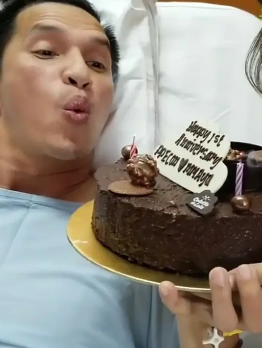 Sambil terbaring di atas kasur rumah sakit, Econ pun meniupkan lilin yang terdapat di atas kue coklat yang bertuliskan “Happy 1st Anniversary PP Econ dan MM Ayu”. Rona bahagia tentunya terlukis di wajah Econ. (Instagram/edison_wardhana)