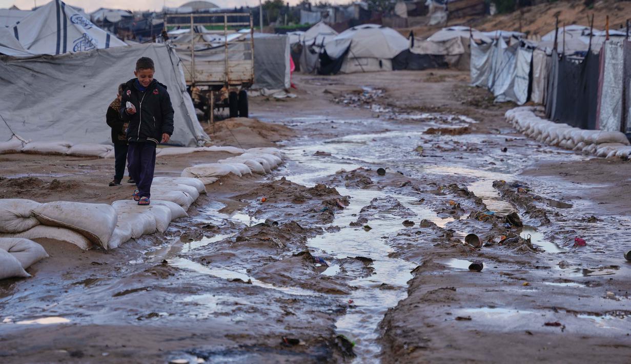 Anak-anak pengungsi Palestina berjalan melewati genangan lumpur setelah hujan deras melanda di sebuah kamp darurat untuk pengungsi, Zawaida, Jalur Gaza tengah, Rabu 17 Desember 2025. Hujan deras dan badai musim dingin Byron yang terjadi sejak pertengahan Desember 2025 telah menyebabkan banjir di berbagai kamp pengungsian yang ada di Jalur Gaza, termasuk di wilayah tengah seperti Zawaida. (AP Photo/Abdel Kareem Hana)
