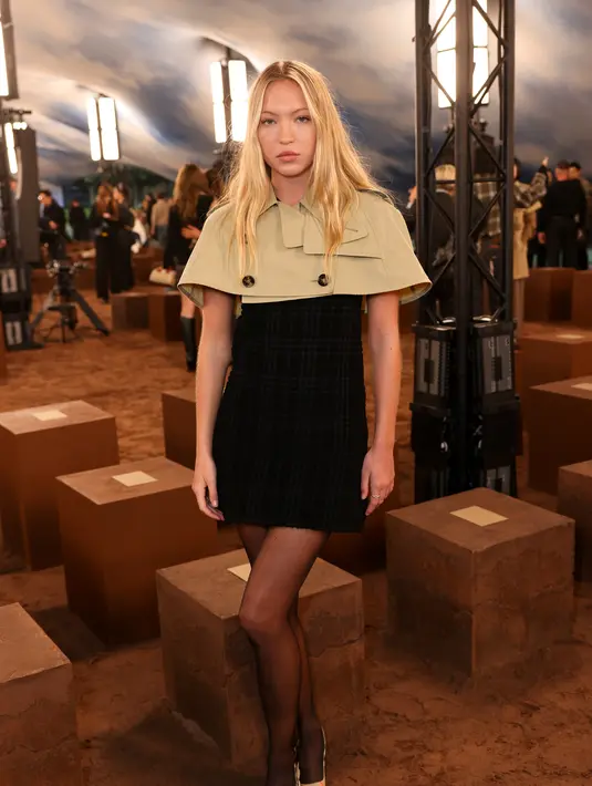Lila Moss tampil playfull dengan mini dress hitam berbalut cropped outer berkancing ganda. Ia juga kenakan stocking hitam dan heels Burberry [Burberry]